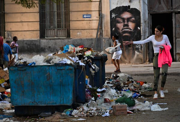 Contenedores azules desbordados y basura cubren una calle. Una persona sostiene una prenda rosa neón, con el brazo extendido, cerca de un edificio con un mural.