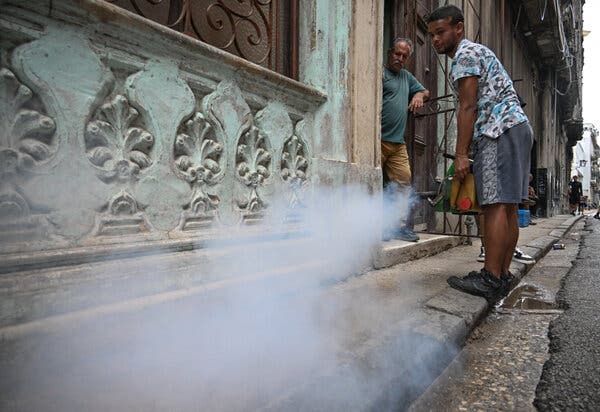 Una densa nube de humo blanco se eleva desde el borde de una calle, donde una persona sostiene un dispositivo amarillo y verde. Cerca, otra persona observa desde la entrada de una casa.