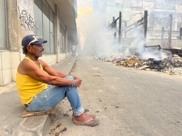 Una persona con un top amarillo, jeans zules y sandalias está sentanda en el bordillo de una acera. Se ve basura humeante.
